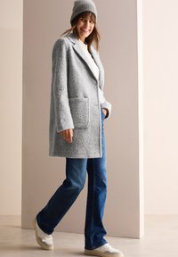 Grått fluffigt ullkappor med en urklippt krage, två framfickor, matchat med blå jeans och vita sneakers, toppat med en grå stickad mössa.