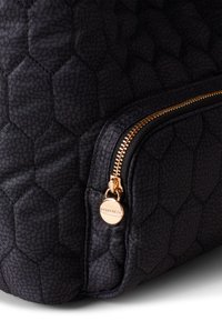 Quilted black fabric backpack with a hexagonal pattern, featuring a gold zipper and round pull tab with engraved logo detail.