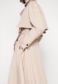 Femme portant un trench-coat beige avec ceinture à la taille et main dans la poche, vue de profil sur un fond uni.
