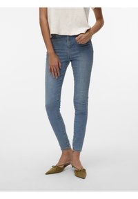 Femme portant un jean skinny bleu clair, un haut court beige et des mules pointues vert olive avec des détails en forme de nœud, debout devant un fond blanc.