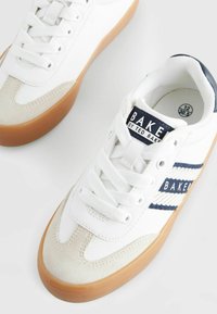 Sneaker blanche avec un embout en daim, des accents bleu marine et blancs rayés, et une semelle en caoutchouc marron. Comprend un col rembourré et des lacets.