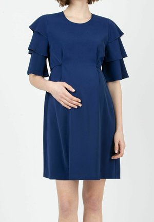 Mujer vestida con un vestido azul marino hasta la rodilla, con mangas cortas de volantes en capas, de pie con una mano apoyada en el abdomen.