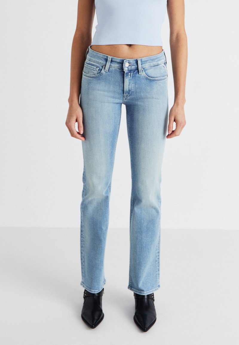 Femme portant un jean droit bleu clair, un crop top blanc et des bottines noires à bout pointu, debout devant un fond uni.