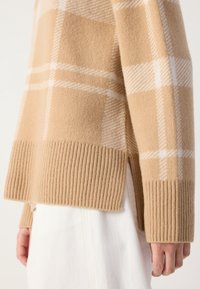 Pull en laine à carreaux beige et blanc avec des poignets et un ourlet côtelés, porté par-dessus une jupe blanche, montrant une fente latérale.