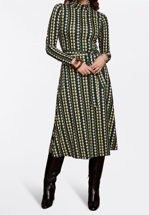Femme portant une robe midi à manches longues avec un motif géométrique vert, blanc et noir, associée à des bottes noires montantes jusqu'aux genoux et des bracelets aux poignets.