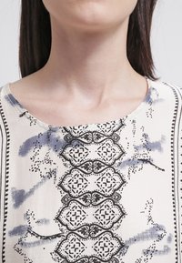 Krämfärgad topp i tyg med rund halsringning, intrikata svarta och blå mönster samt dekorativa kanter längs sidorna.