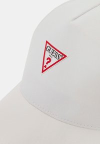 White cotton cap featuring a triangular red logo patch with a white question mark and "GUESS U.S.A." text. Smooth texture, structured design.