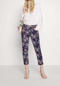 Blouse blanche à manches longues associée à un pantalon bleu marine à motifs floraux et des sandales à talons hauts beige à brides, avec une ceinture et un sac à main dorés.