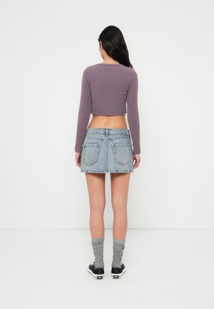 Kobieta z długimi ciemnymi włosami, ubrana w fioletowy, długi crop top, niebieską dżinsową mini spódniczkę, szare pomięte skarpetki i czarne trampki, stojąca tyłem.