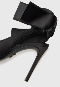 Crna satenirana cipelica na visokom petu sa šiljastim prstom i velikim detaljem od vrpce oko gležnja, sa sjajnom stiletto petom.