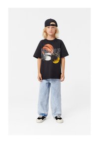 Crna oversized majica s koloristom sportskim grafičkim uzorkom; svijetloplave široke traperice; crna kapa; crno-bijele tenisice. Jednostavna pozadina.