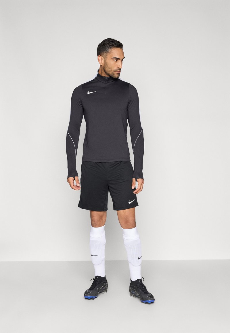Schwarzes langärmliges Sportoberteil mit halbhohem Reißverschluss, kombiniert mit schwarzen Shorts, weißen Socken und schwarzen Fußballschuhen mit blauen Akzenten. Nike-Logo sichtbar.