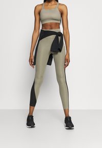 Světle olivový crop top a legíny s vysokým pasem, s černými akcenty v pase a po stranách. Modelka má na sobě černé sportovní boty.