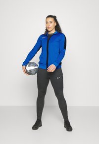 Blauwe sportjack met rits en zwarte accenten, gecombineerd met zwarte leggings met geometrische patronen. Houdt een zilveren voetbal vast.