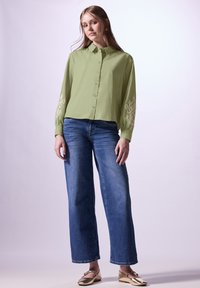 Camicia verde oliva con bottoni e dettagli ricamati sulle maniche, abbinata a jeans a gamba larga blu e ballerine metallizzate dorate.