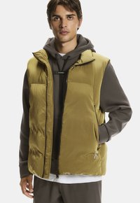 Junger Mann mit olivgrünem Puffy-Weste über grauem Hoodie, der mit einer Hand in der Weste- Tasche vor einem schlichten Hintergrund steht.