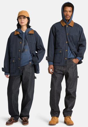 Kaksi mallia, jotka pukeutuvat tummansinisiin takkeihin, joissa on ruskeat kaulukset, siniset hupulliset paidat, tummat farkut ja saappaat; yksi mallista käyttää myös beigeä lippalakkia.