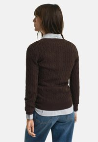 Brauner kabelgestrickter Pullover mit gerippten Bündchen und Saum, über einem hellblauen und weißen gestreiften Hemd mit Kragen getragen, kombiniert mit blauen Jeans.