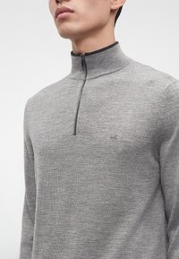 Homme portant un pull en tricot gris clair avec une demi-fermeture éclair bordée de noir et un petit logo "MK" sur le côté gauche de la poitrine.