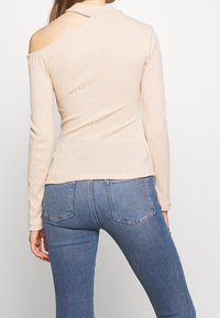 Vaalean beige ribbineuleinen pitkähihainen paita, jossa on avoin olkapään muotoilu, korkea kaulus ja istuva malli. Yhdistettynä sinisiin keski vyötäröisiin farkkuihin.