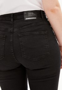 Jeans de mezclilla negra de cintura alta, con dos bolsillos traseros y una etiqueta de tela rectangular con instrucciones de cuidado. Textura suave.