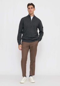 Uomo che indossa un maglione con zip grigio scuro, pantaloni marroni e scarpe da ginnastica bianche, in piedi davanti a uno sfondo bianco uniforme.