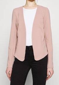 Blazer rose clair en tissu doux, avec un design sans col, manches longues et ourlet arrondi. Associé à une chemise blanche et un pantalon noir.