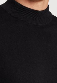 Zwarte coltrui met een geribde kraag en een zachte, gebreide textuur. De close-up benadrukt de warmte en zachtheid van de stof.