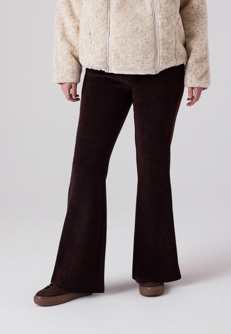 Braune Cordhosen mit ausgestelltem Bein und strukturierter Oberfläche, kombiniert mit einer cremefarbigen Fleecejacke. Die Schuhe sind braun mit strukturierten Akzenten.