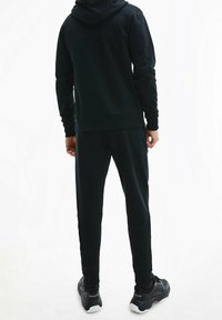 Chándal deportivo negro que incluye sudadera con capucha y pantalones ajustados. Tejido suave con puños de canalé, cinturilla elástica y detalles de logotipo.