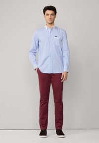 Camisa abotonada de color azul claro con finas rayas verticales, bolsillo en el pecho; combinada con pantalones slim-fit color burdeos y zapatillas oscuras.