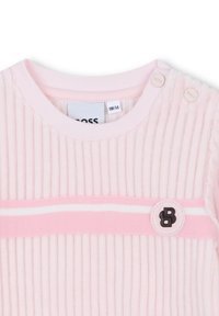 Haut en tissu côtelé rose clair avec une bande horizontale blanche et un accent de logo rond. Comprend deux fermetures à bouton-pression sur une épaule.