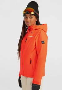Feloranje waterdichte jas met capuchon, ritszakken en een zwart logo. Gepaard met zwarte handschoenen en een skibril. Glad oppervlak.