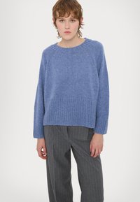 Maglione blu a maglia con scollo rotondo, orlo e polsini a coste, caratterizzato da una vestibilità ampia e un tessuto a trama, indossato con pantaloni grigi a righe.