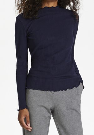 Femme portant un shirt à manches longues côtelé bleu marine avec des bords festonnés et un pantalon gris clair, debout avec une main dans la poche.