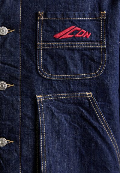 Denim shirt featuring a dark blue color, yellow stitching, a chest pocket with red logo embroidery, and metal snap buttons.