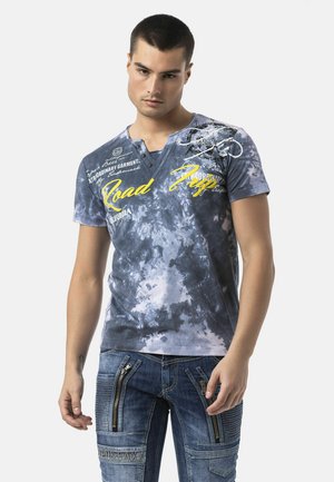 Junger Mann mit kurzen dunklen Haaren, der ein blaues Batik-Grafik-T-Shirt und blaue Jeans mit mehreren Reißverschlüssen und Taschen trägt.