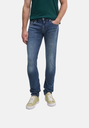 Jeans de mezclilla azul con un corte slim, que presentan un lavado medio, un ligero desvanecimiento y un diseño clásico de cinco bolsillos, combinados con zapatillas de color verde claro.
