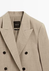 Beige dubbelknäppt blazer med slagkrage och svarta knappar, märkt Massimo Dutti, visad mot en vit bakgrund.