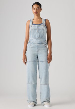 Femme portant une salopette en denim bleu clair et des baskets, debout face à un fond gris uni.