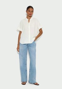 Una donna è in piedi contro uno sfondo neutro, indossa una camicia bianca a maniche corte, jeans larghi di colore blu chiaro e sandali neri.