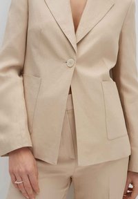 Blazer in lino beige con chiusura a un bottone, rever a pugno e due tasche frontali. Tessuto liscio, vestibilità sartoriale, pantaloni beige abbinati.