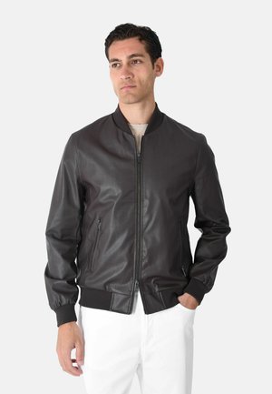 Giovane uomo che indossa una giacca bomber in pelle nera con zip, polsini e colletto a coste, abbinata a pantaloni bianchi, in piedi davanti a uno sfondo neutro.