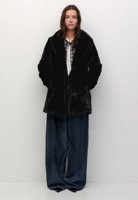 Vrouw in een zwarte nep-bontjas, geruite shirt met een stropdas en wijde donkerblauwe jeans, staand tegen een effen witte achtergrond.