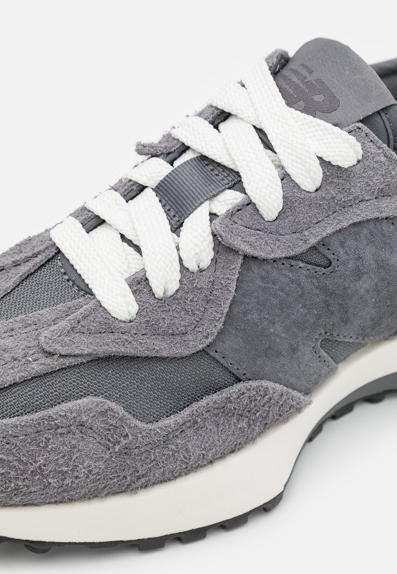 Zapatilla gris que presenta una combinación de materiales de ante y tela, superficie texturizada, cordones blancos prominentes y una gruesa suela de goma negra.