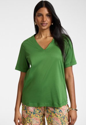 Kobieta z długimi czarnymi włosami nosi zielony top z krótkim rękawem i dekoltem w serek oraz kolorowe spodnie w kwiatowy wzór, uzupełnione złotymi kolczykami i bransoletką.