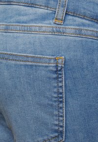 Jeans de ganga azul com um bolso traseiro, apresentando costura amarela e um acabamento texturizado clássico. O tecido tem uma aparência desgastada.
