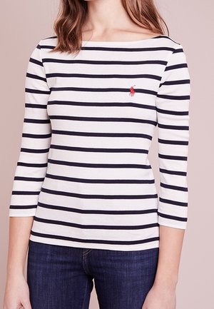 Kvinde iført en hvid og marineblå stribet langærmet bluse med en rød broderet logo på brystet, kombineret med mørkeblå jeans.