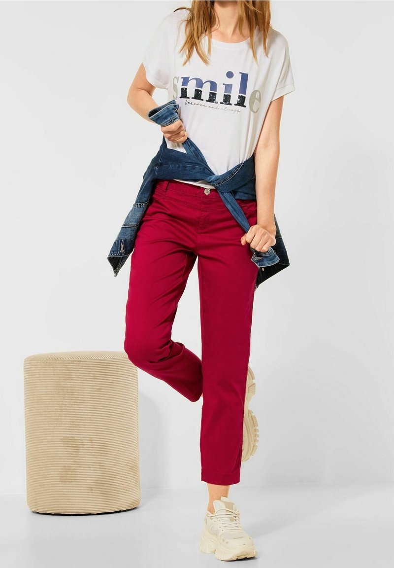 Red pants with side pockets, paired with a white t-shirt featuring a printed design. A denim jacket is tied around the waist. Beige stool in background.