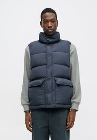 Chaleco acolchado azul marino con un cuello alto, cierre de botones, dos bolsillos frontales y una textura acolchada. Lleva puesto una camiseta de manga larga gris.